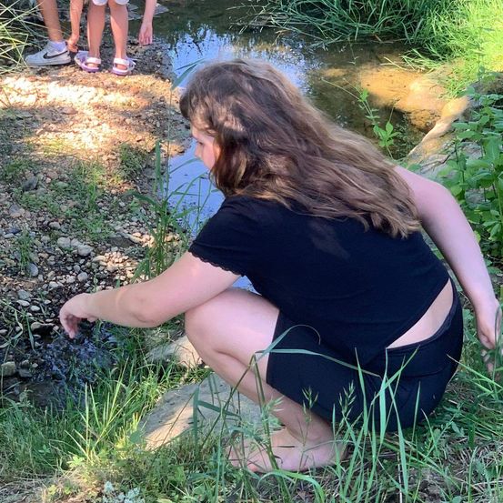 Eine junge Frau hockt sich neben einem kleinen Bach und untersucht das Wasser, während zwei Kinder hinter ihr stehen.