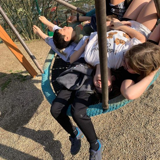 Vier Kinder liegen auf einer Schaukel in einem Außenspielplatz, wobei ein Mädchen eine Brille und Turnschuhe trägt.