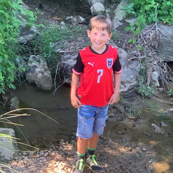 Ein junger Junge in einem roten Fußballtrikot mit der Nummer 7 steht in einem kleinen Bach. Er trägt Jeansshorts und Turnschuhe. Hinter ihm befinden sich Steine und grüne Pflanzen.