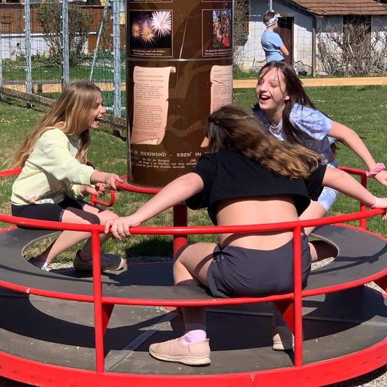 Drei Mädchen spielen auf einem Karussell in einem Park. Sie lachen und haben Spaß. Das Mädchen in der Mitte schiebt die anderen.