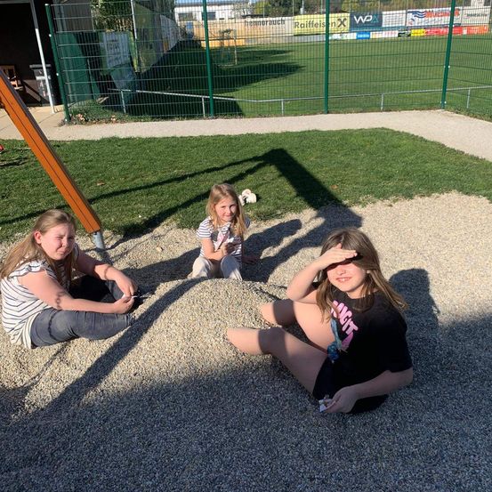 Drei Mädchen sitzen auf dem Kies in einem Freispielplatzbereich, ein Mädchen hält etwas in der Hand. Hinter ihnen steht ein grüner Zaun.