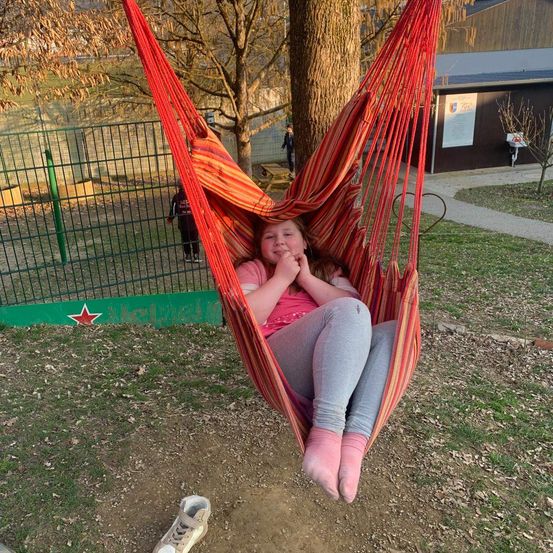 Ein junges Mädchen in einem roten Hängestuhl lächelt in die Kamera, während es in einem Park sitzt, mit einem Schuh in der Nähe.