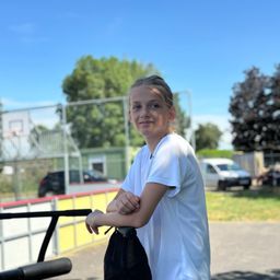 Ein junges Mädchen in einem weißen T-Shirt steht in einem Außenbereich, vielleicht einem Park, mit einem Basketballplatz und Bäumen im Hintergrund.