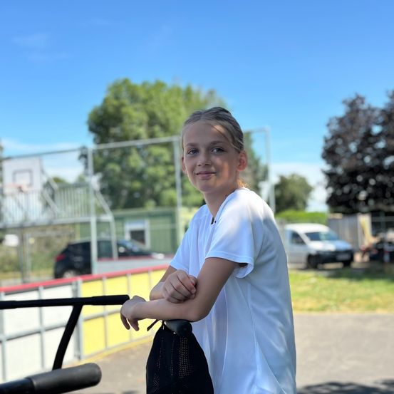 Ein junges Mädchen in einem weißen T-Shirt steht in einem Außenbereich, vielleicht einem Park, mit einem Basketballplatz und Bäumen im Hintergrund.