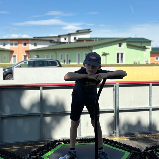Ein Junge in einer Mütze und Turnschuhen steht auf einem Trampolin und schaut nach unten. Hinter ihm ein Zaun und ein Gebäude mit grünen Fenstern.