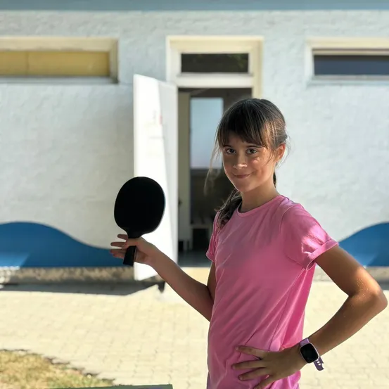 Ein junges Mädchen posiert draußen, hält einen schwarzen Tischtennisschläger in der Hand, lächelt und steht vor einem Gebäude mit weißen Wänden und blauer Einfassung.