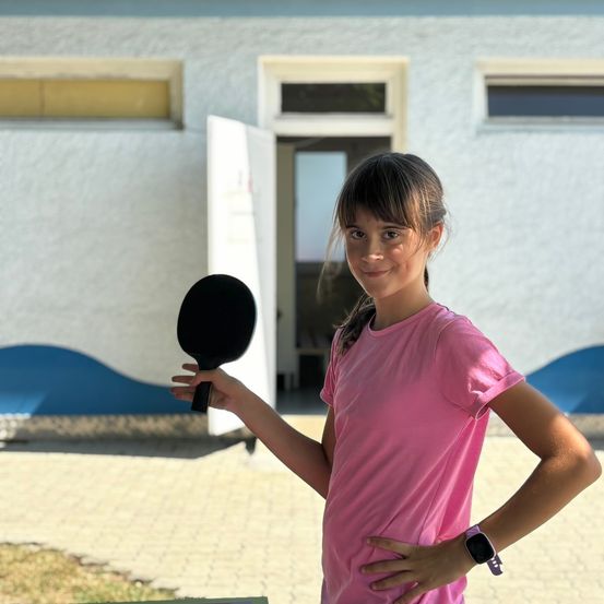 Ein junges Mädchen posiert draußen, hält einen schwarzen Tischtennisschläger in der Hand, lächelt und steht vor einem Gebäude mit weißen Wänden und blauer Einfassung.