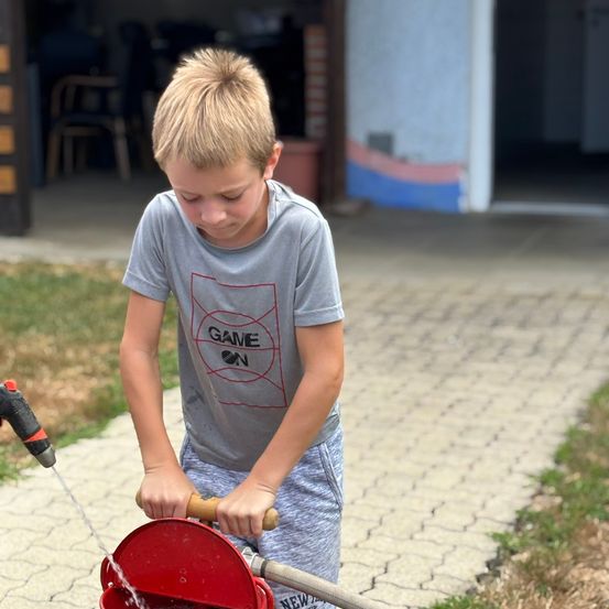 Ein Junge steht draußen, hält einen Schlauch in der Hand und füllt einen roten Eimer mit Wasser. Er trägt ein graues T-Shirt mit rotem Aufdruck.