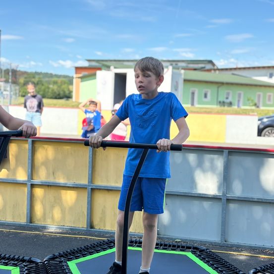 Ein junger Junge in einem blauen T-Shirt steht auf einem Trampolin und hält einen Griff. Andere Kinder sind im Hintergrund.