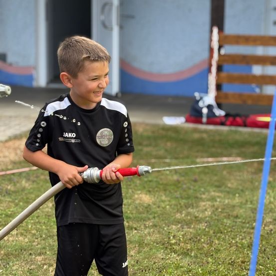 Ein junger Junge hält einen Wasserschlauch und sprüht Wasser nach draußen. Er trägt ein schwarzes und weißes Fußballtrikot.