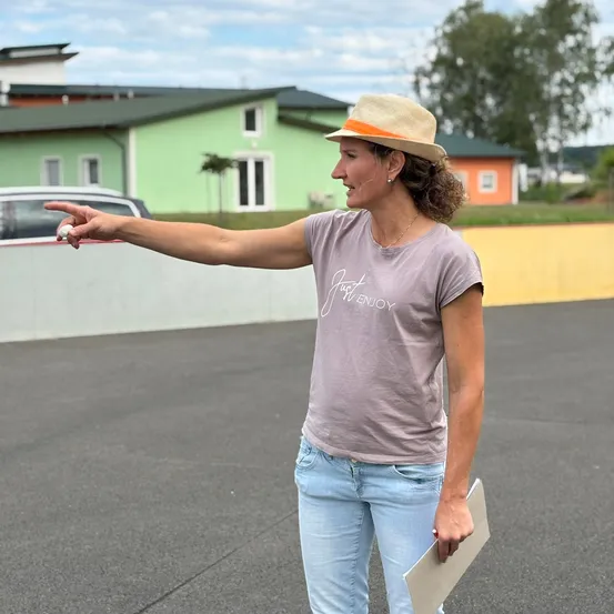 Eine Frau in einem Hut zeigt mit ihrem Finger draußen auf einem Parkplatz. Ein grünes Gebäude und ein Auto sind hinter ihr.