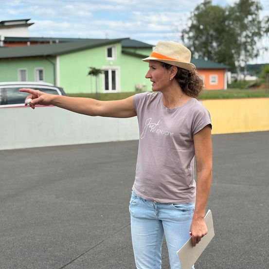 Eine Frau in einem Hut zeigt mit ihrem Finger draußen auf einem Parkplatz. Ein grünes Gebäude und ein Auto sind hinter ihr.