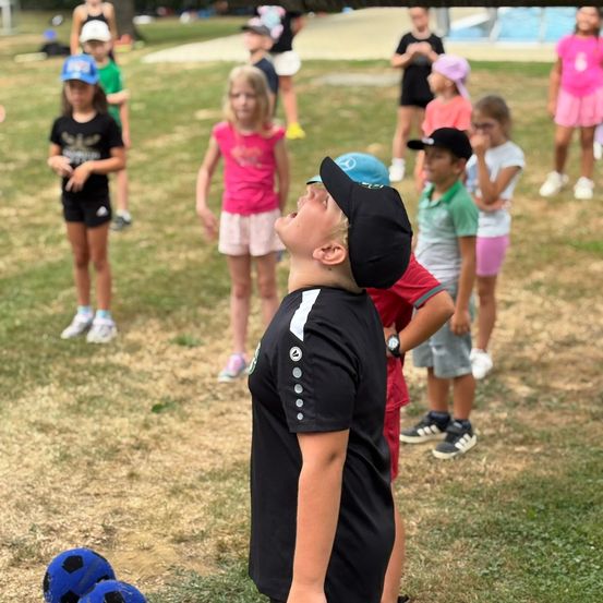 Eine Gruppe von Kindern in Outdoor-Sportbekleidung, einige mit Mützen und Turnschuhen, üben Fußball. Zwei Jungen schauen nach oben, während andere herumstehen.