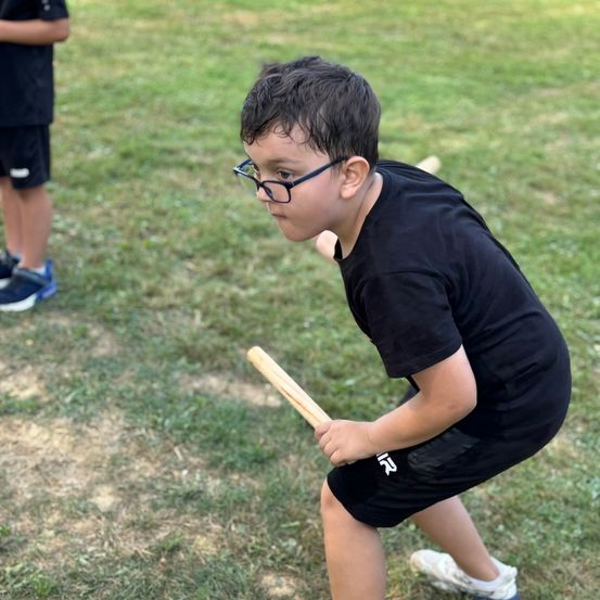 Ein Junge mit Brille schwingt einen Holzschläger auf einem Rasen, ein anderer Junge steht hinter ihm.