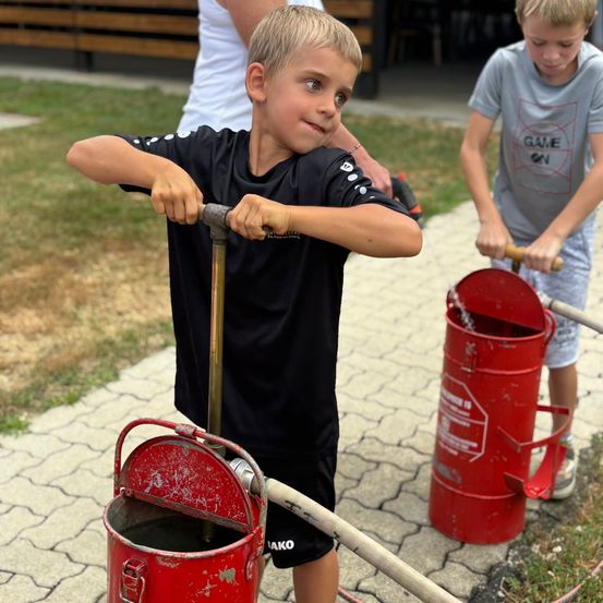 Zwei junge Jungen sind draußen und benutzen rote Feuerlöscher. Ein Junge hält einen Schlauch und der andere benutzt eine Pumpe.