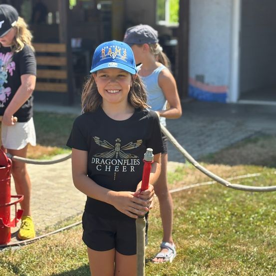 Ein Mädchen in einem Dragonflies Cheer T-Shirt steht draußen und hält einen Feuerwehrschlauch, lächelt, während zwei andere Mädchen im Hintergrund stehen.