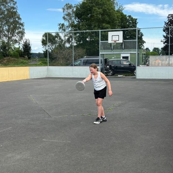 Ein Mädchen spielt Frisbee auf einem Freiluftplatz mit einem Basketballkorb im Hintergrund, unter einem blauen Himmel.