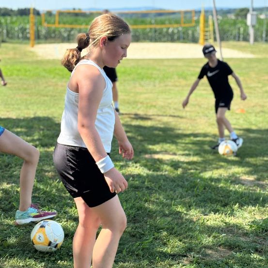 Ein Mädchen in einem weißen Tanktop und schwarzen Shorts spielt mit anderen auf einem Rasenfeld Fußball. Ein Junge in Schwarz läuft auf den Ball zu.