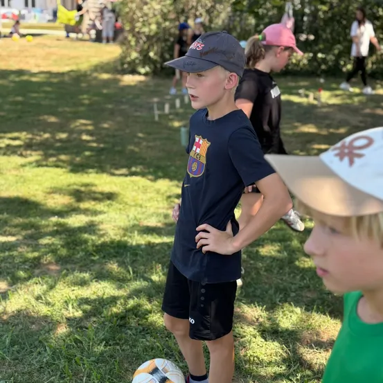 Ein Junge mit einem schwarzen Hut und einem Barcelona-T-Shirt steht neben einem Fußball auf einem Rasenfeld. Dahinter spielen andere Kinder Fußball.