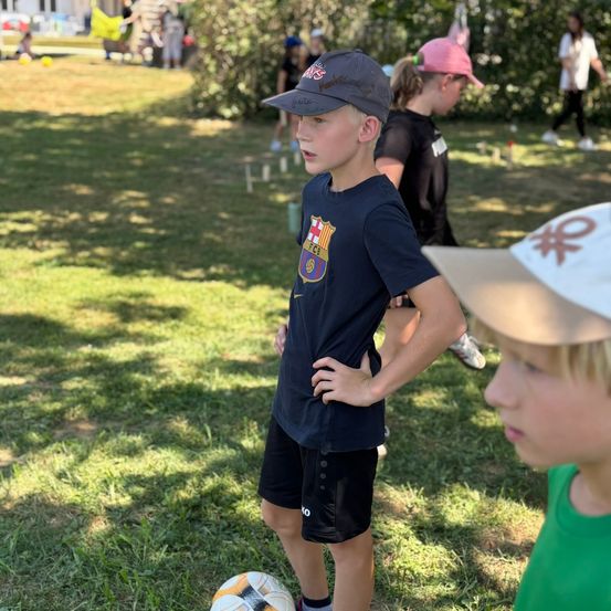 Ein Junge mit einem schwarzen Hut und einem Barcelona-T-Shirt steht neben einem Fußball auf einem Rasenfeld. Dahinter spielen andere Kinder Fußball.