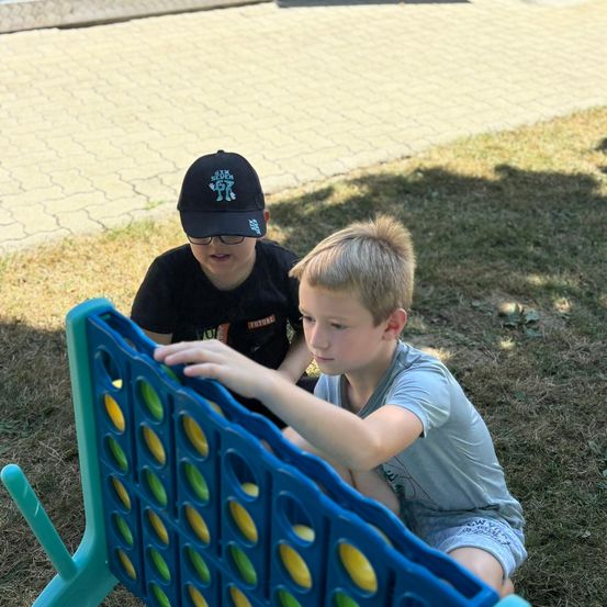 Zwei Jungen spielen ein Spiel von Vier Gewinnt auf einem Grasfeld, einer trägt eine Baseballkappe.