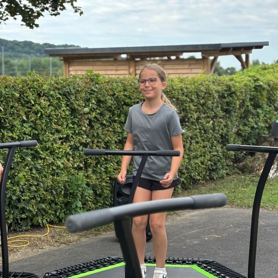 Ein junges Mädchen mit Brille steht lächelnd auf einem Trampolin, mit einem Busch und einer Holzstruktur im Hintergrund.