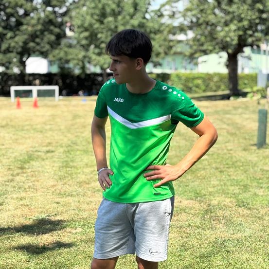 Ein junger Mann steht auf einem Rasenfeld und trägt ein grünes T-Shirt und graue Shorts. Er hat die Hand auf der Hüfte. Im Hintergrund ist ein Fußballtor zu sehen.