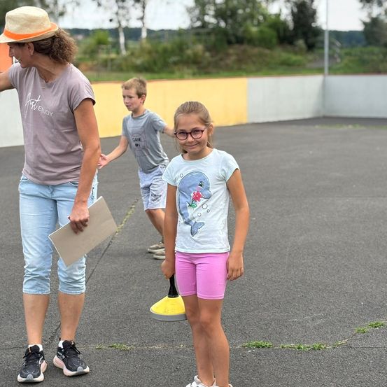 Eine Frau und zwei Kinder stehen auf einem Parkplatz. Die Frau hält ein Papier, und ein Mädchen mit Brille schaut in die Kamera. Hinter ihnen geht ein Junge mit den Händen hinter dem Rücken.