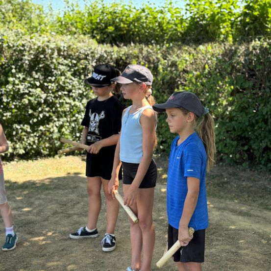Drei Kinder in einem Park, zwei halten Stöcke, eines in einem blauen Shirt, ein anderes in einem weißen Tanktop, alle tragen Mützen. Dahinter steht ein Busch.