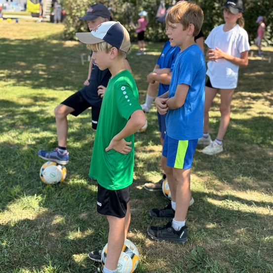 Mehrere Kinder stehen auf einem Grasfeld und bereiten sich auf ein Fußballspiel vor. Ein Kind in Grün schaut auf seinen Ball hinunter.