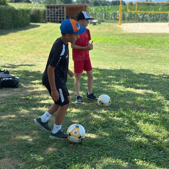 Zwei Jungen spielen Fußball auf einem Grasfeld mit einem Volleyballnetz im Hintergrund. Ein Junge trägt ein schwarzes Shirt, der andere ist in Rot.