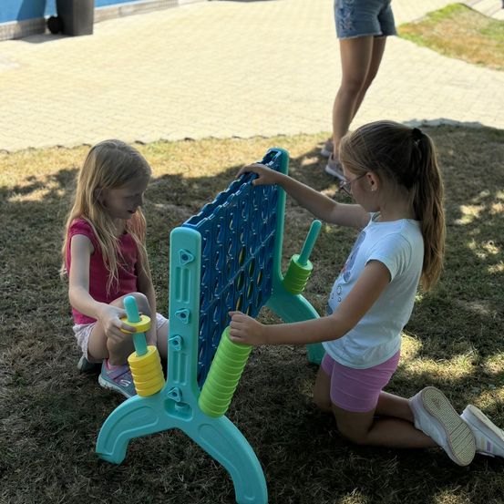 Zwei Mädchen spielen an einem sonnigen Tag. Eines hält einen gelben und grünen Spinner. Das andere trägt eine Brille und hält ein blaues Brett mit Löchern.