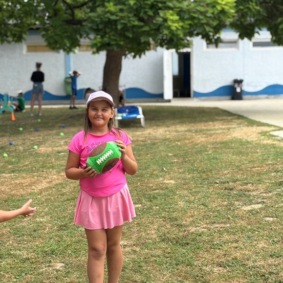 Ein junges Mädchen in einem rosa Shirt und Rock steht auf einem Feld und hält einen Football. Mehrere andere Kinder spielen im Hintergrund in der Nähe eines Gebäudes.