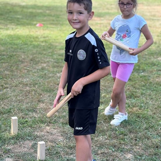 Zwei Kinder spielen mit Holzstäben und kleinen Holzpfosten in einer Grasfläche. Der Junge hält einen Stab, während das Mädchen zwei Stäbe hält.