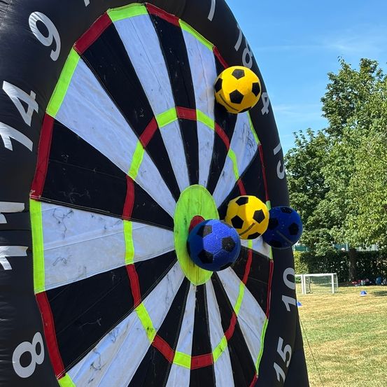 Ein aufblasbares Dartboard mit aufgesteckten Fußballen, die die Zahlen 149, 140, 135, 10 und 8 anzeigen. Es steht auf einem Rasenfeld mit Bäumen und einem Fußballtor im Hintergrund.