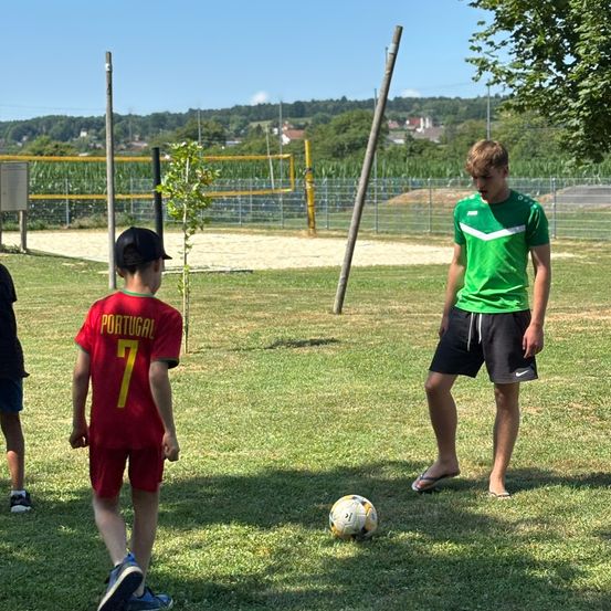 Ein Junge in einem roten Portugal-Trikot steht neben einem Fußball. Ein Erwachsener in Grün steht in der Nähe und schaut zu. Im Hintergrund eine Mauer und ein Volleyballnetz.