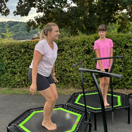 Zwei Frauen stehen auf Trampolins, eine mit den Füßen auf dem Trampolin und die andere am Rand. Sie sind draußen in einem Garten.