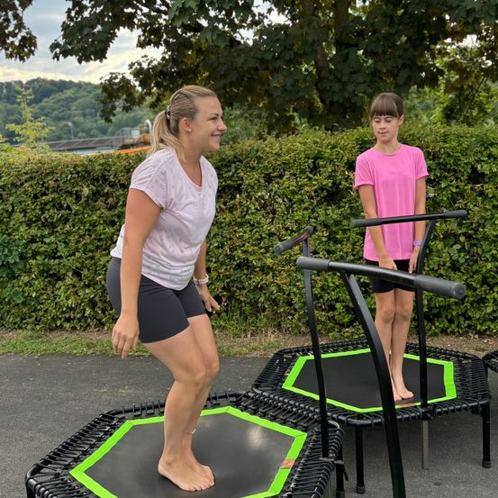 Zwei Frauen stehen auf Trampolins, eine mit den Füßen auf dem Trampolin und die andere am Rand. Sie sind draußen in einem Garten.