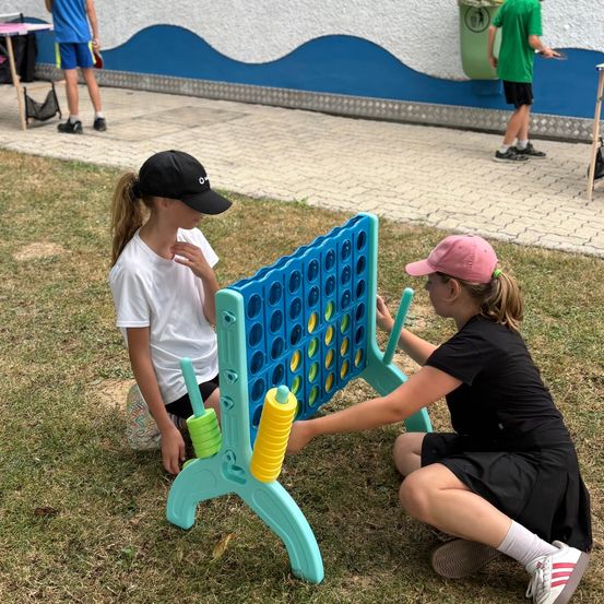 Zwei junge Mädchen spielen an einem sonnigen Tag ein Spiel Connect Four. Eines der Mädchen trägt ein weißes T-Shirt und das andere schwarz. Ein bunter Tisch steht auf dem Rasen.