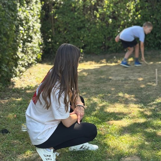 Ein junges Mädchen in einem weißen T-Shirt beobachtet einen Jungen, der auf dem Rasen ein Spiel spielt. Der Junge ist gebeugt und hält einen Stock, vielleicht spielt er Croquet.