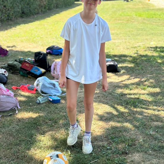 Ein junges Mädchen in einem weißen T-Shirt und Turnschuhen steht auf einem Rasenfeld mit einem Fußball. Mehrere Rucksäcke und Gegenstände sind verstreut.