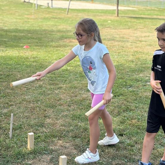 Ein Mädchen mit Brille und einem Delphin-Shirt spielt mit einem Jungen auf einem Grasfeld. Sie halten Stöcke.