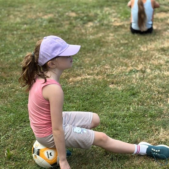 Ein Mädchen sitzt auf einem Fußball, trägt ein rosa T-Shirt und graue Shorts, während ein anderes Mädchen in einem weißen T-Shirt hinter ihr kniet.