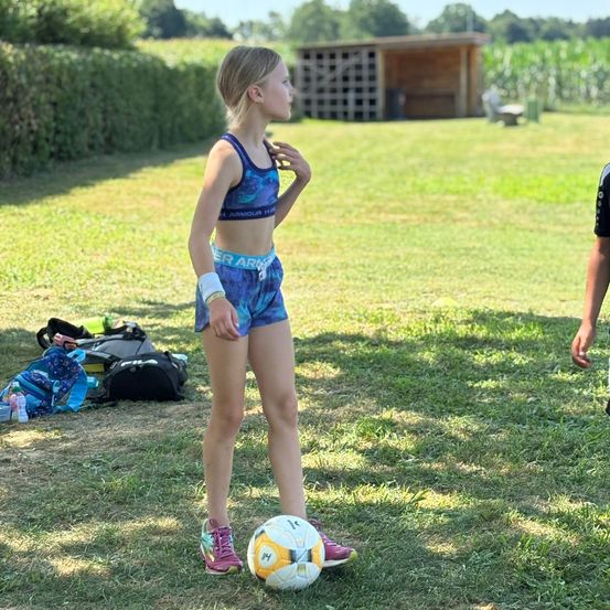 Ein junges Mädchen in Sportkleidung steht auf einem Rasenfeld, schaut zur Seite und hat einen Fußball vor den Füßen.