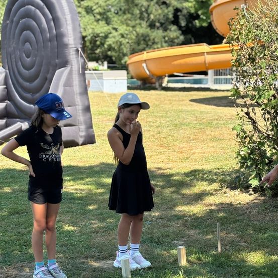 Zwei Mädchen stehen in der Nähe eines Spielplatzes mit einer Rutsche und einer riesigen Schnecke. Ein Mädchen hat eine Kappe, und das andere hat ein Kleid. Sie scheinen ein Spiel zu spielen.