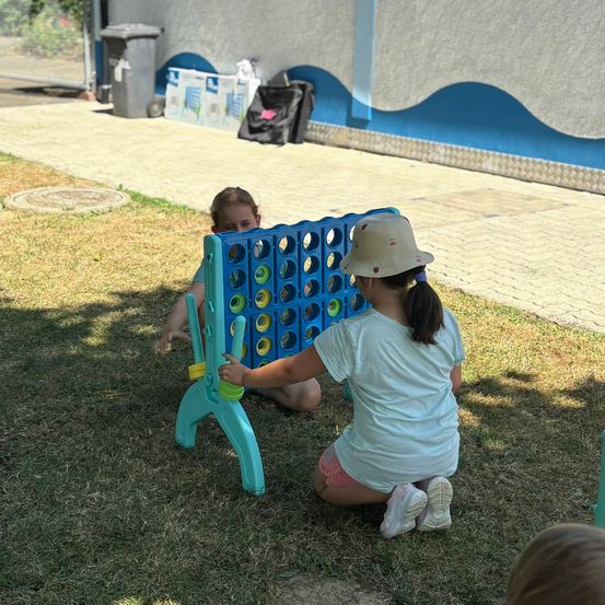 Zwei Mädchen spielen eine Partie Connect Four auf einem Rasen. Eines trägt einen Hut und die andere ein Armband.