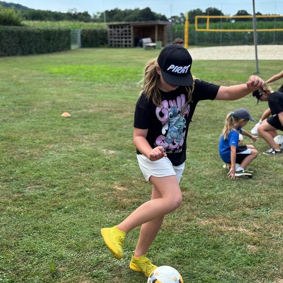 Ein junges Mädchen in einem schwarzen Shirt mit Piratenhut tritt einen Fußball auf einem Rasenfeld. Andere Kinder sitzen im Hintergrund.