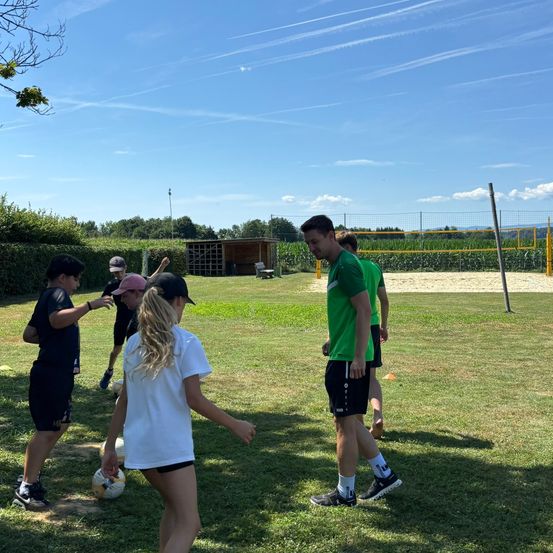 Mehrere Kinder und ein Trainer üben Fußball auf einem Feld mit einem Netz und einem Gebäude in der Ferne. Sie tragen Sportkleidung und halten Fußballbälle.