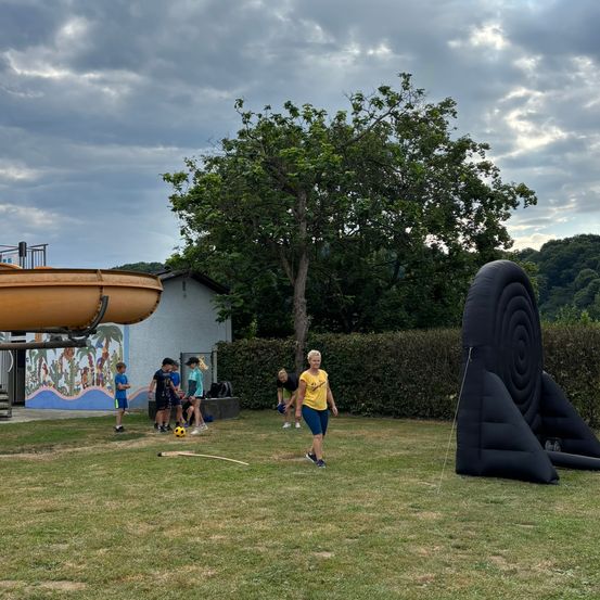 Eine Gruppe von Kindern und eine erwachsene Frau spielen ein Spiel auf einem Grasfeld. Es gibt eine Wasserrutsche, ein großes aufblasbares Objekt und ein Gebäude mit Wandkunst im Hintergrund.