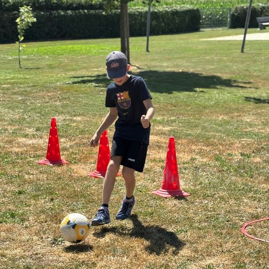 Ein Junge spielt Fußball auf einem Rasenfeld mit einem Fußball, trägt einen Hut und Turnschuhe.